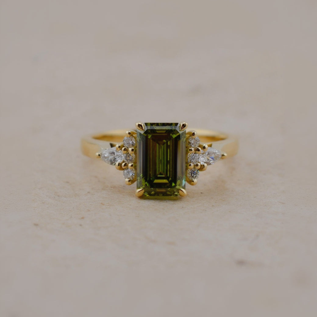 A yellow gold ring featuring a rectangular emerald-cut olive green gemstone center, accented by small clear stones on either side of the band, set against a neutral beige background.