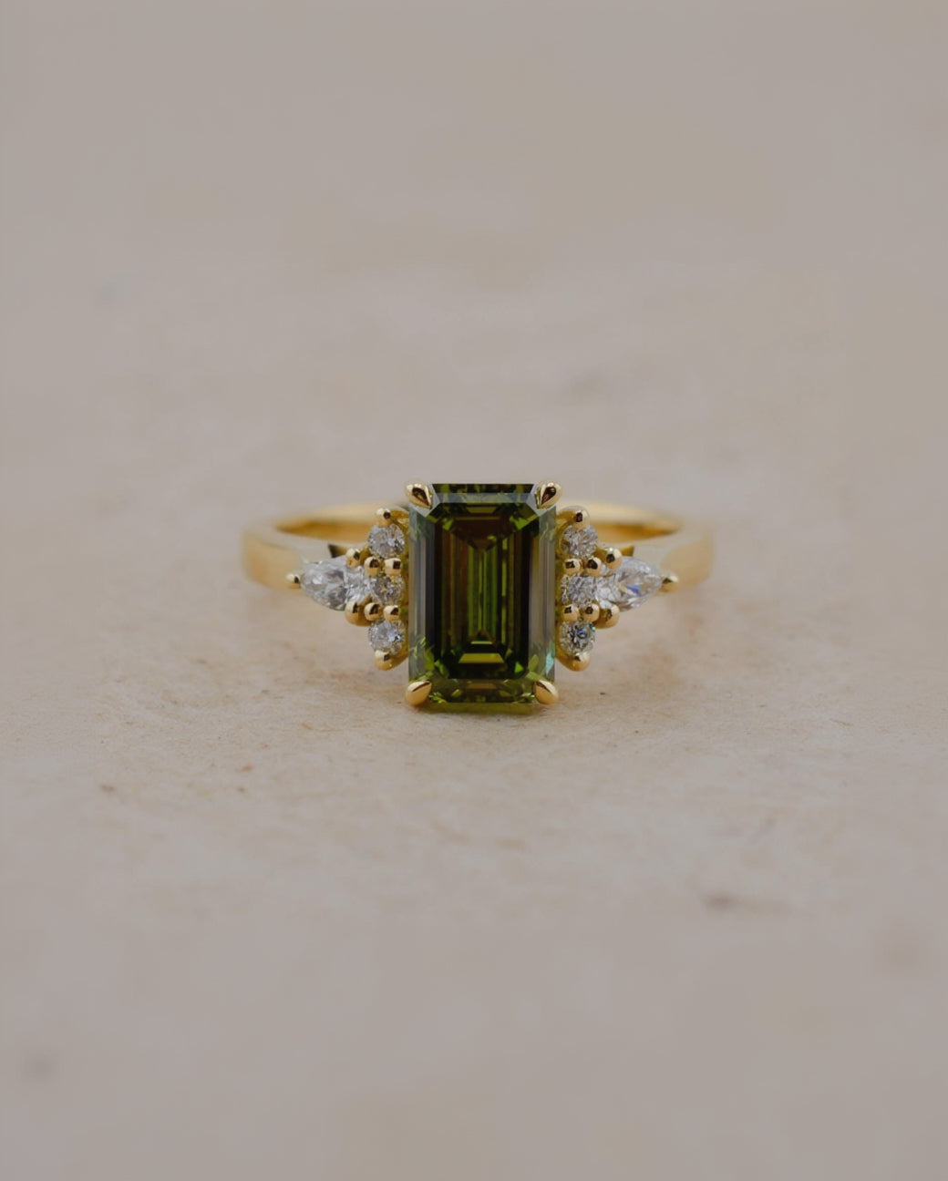 A yellow gold ring featuring a rectangular emerald-cut olive green gemstone center, accented by small clear stones on either side of the band, set against a neutral beige background.