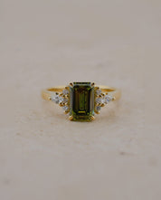 A yellow gold ring featuring a rectangular emerald-cut olive green gemstone center, accented by small clear stones on either side of the band, set against a neutral beige background.