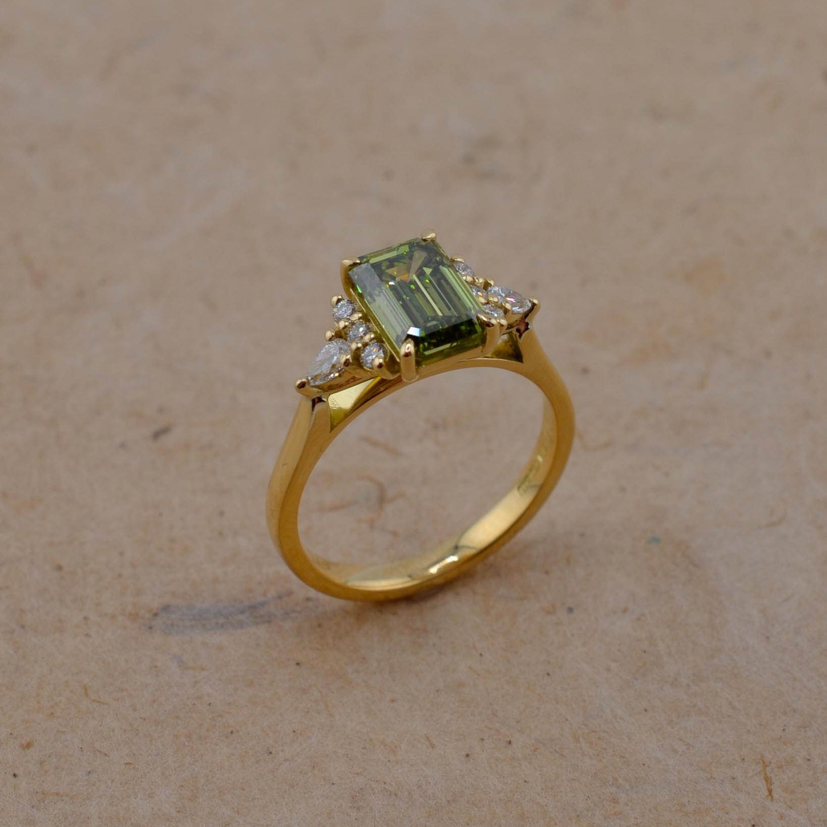 A yellow gold ring featuring a rectangular emerald-cut green gemstone and small clear accent stones, displayed on a textured beige surface.
