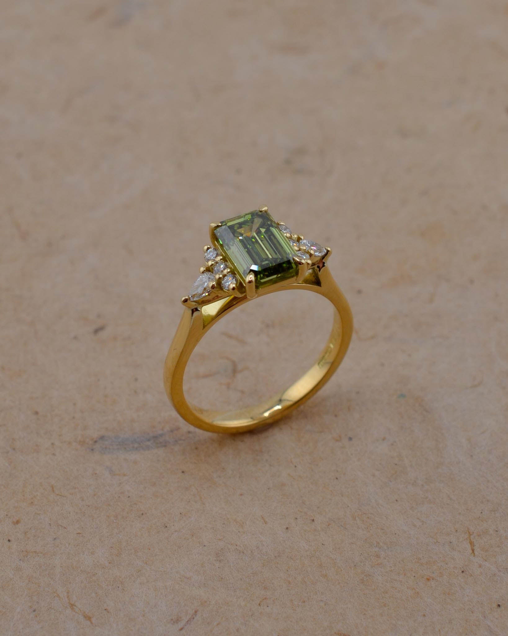 A yellow gold ring featuring a rectangular emerald-cut green gemstone and small clear accent stones, displayed on a textured beige surface.