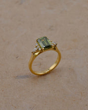 A yellow gold ring featuring a rectangular emerald-cut green gemstone and small clear accent stones, displayed on a textured beige surface.