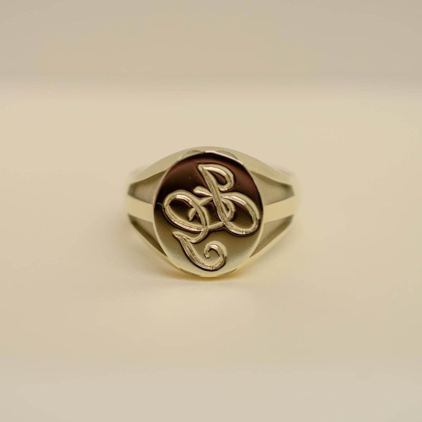 A gold signet ring with a polished oval face featuring an interlocking monogram design, set against a plain light beige background.