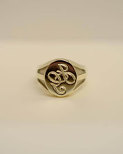 A gold signet ring with a polished oval face featuring an interlocking monogram design, set against a plain light beige background.