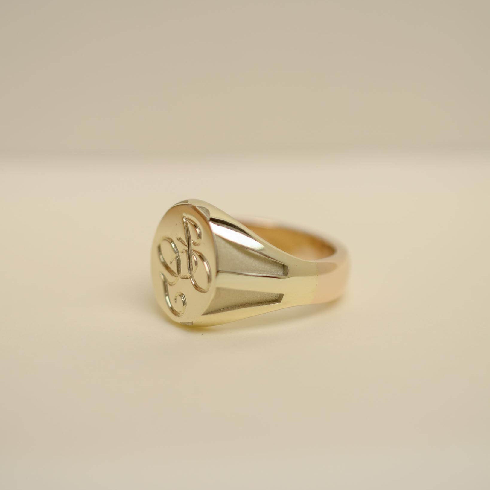 A polished yellow gold signet ring with an engraved floral motif on its oval face, resting on a neutral beige surface.