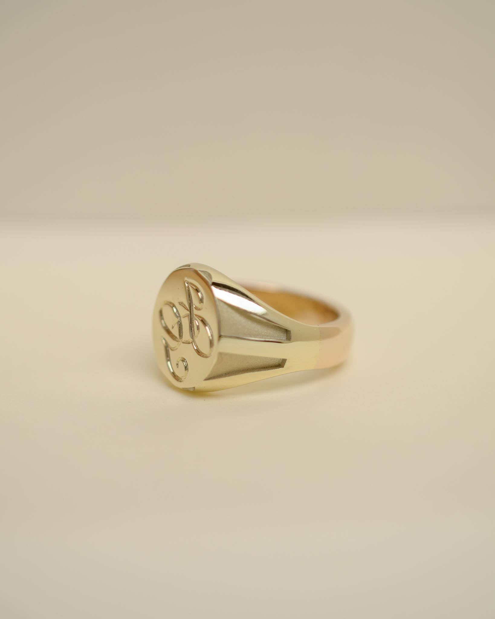 A polished yellow gold signet ring with an engraved floral motif on its oval face, resting on a neutral beige surface.