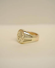 A polished yellow gold signet ring with an engraved floral motif on its oval face, resting on a neutral beige surface.