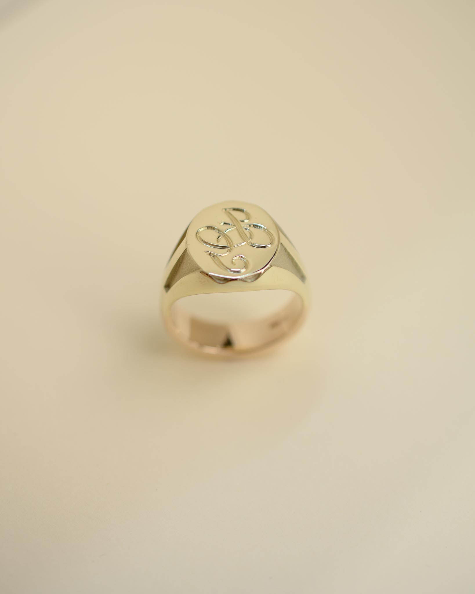 A polished gold signet ring with an engraved monogram on a smooth, light beige background.