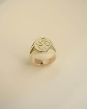 A polished gold signet ring with an engraved monogram on a smooth, light beige background.