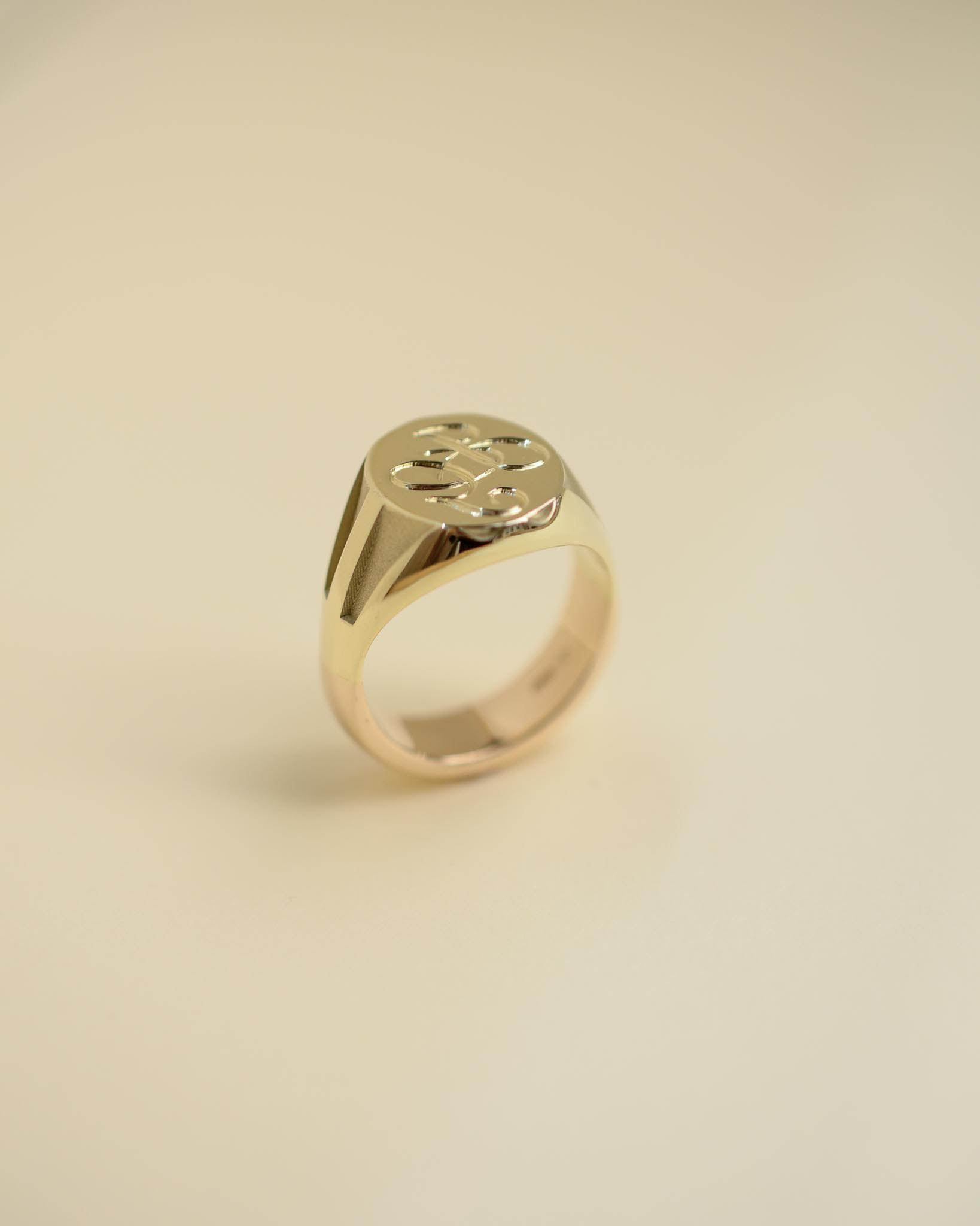 A polished gold signet ring with a stylized cutout monogram on the face, displayed on a clean, solid cream-colored background.