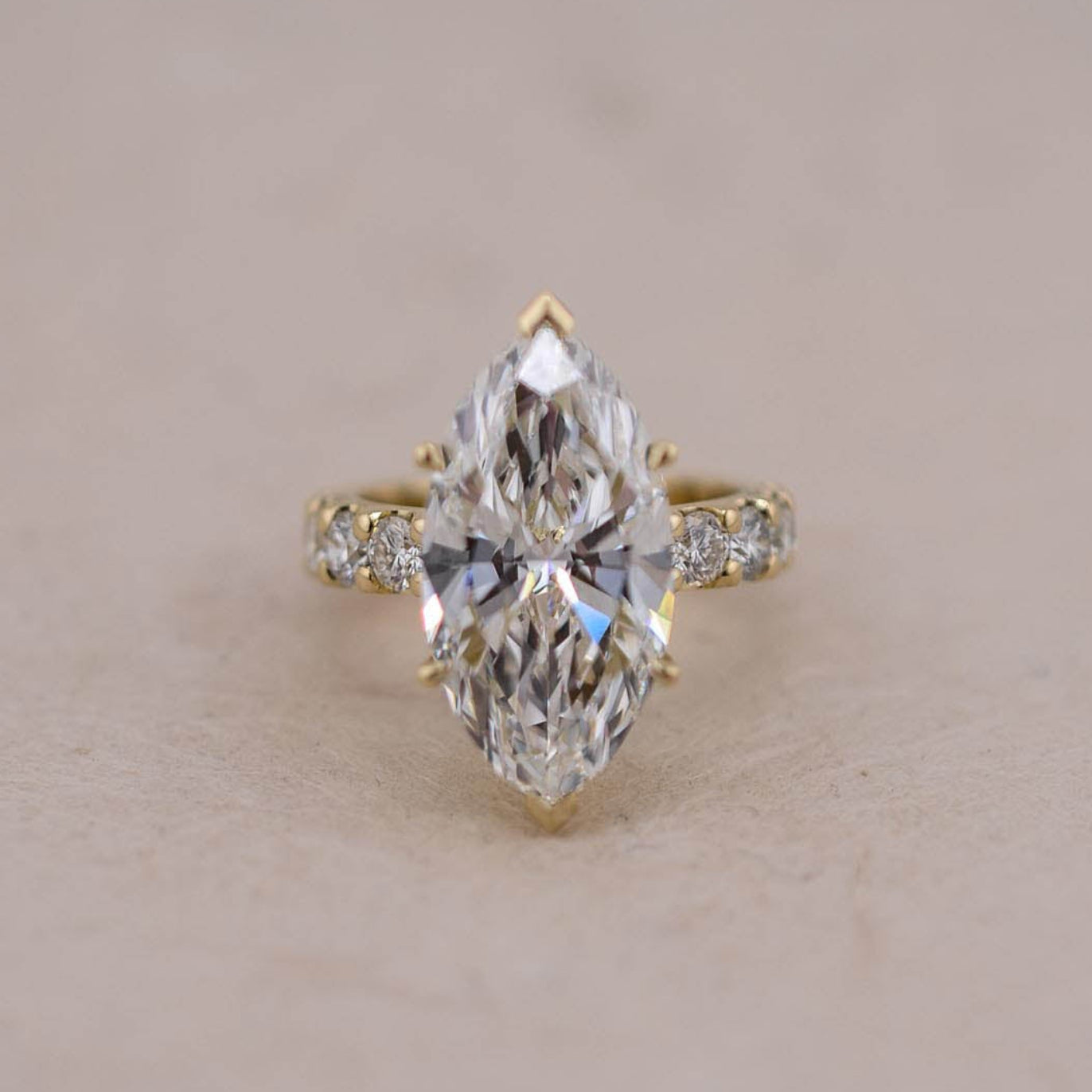 A yellow gold ring featuring a marquise-cut white diamond in a prong setting, accented with small round diamonds on the band, set against a plain beige background.