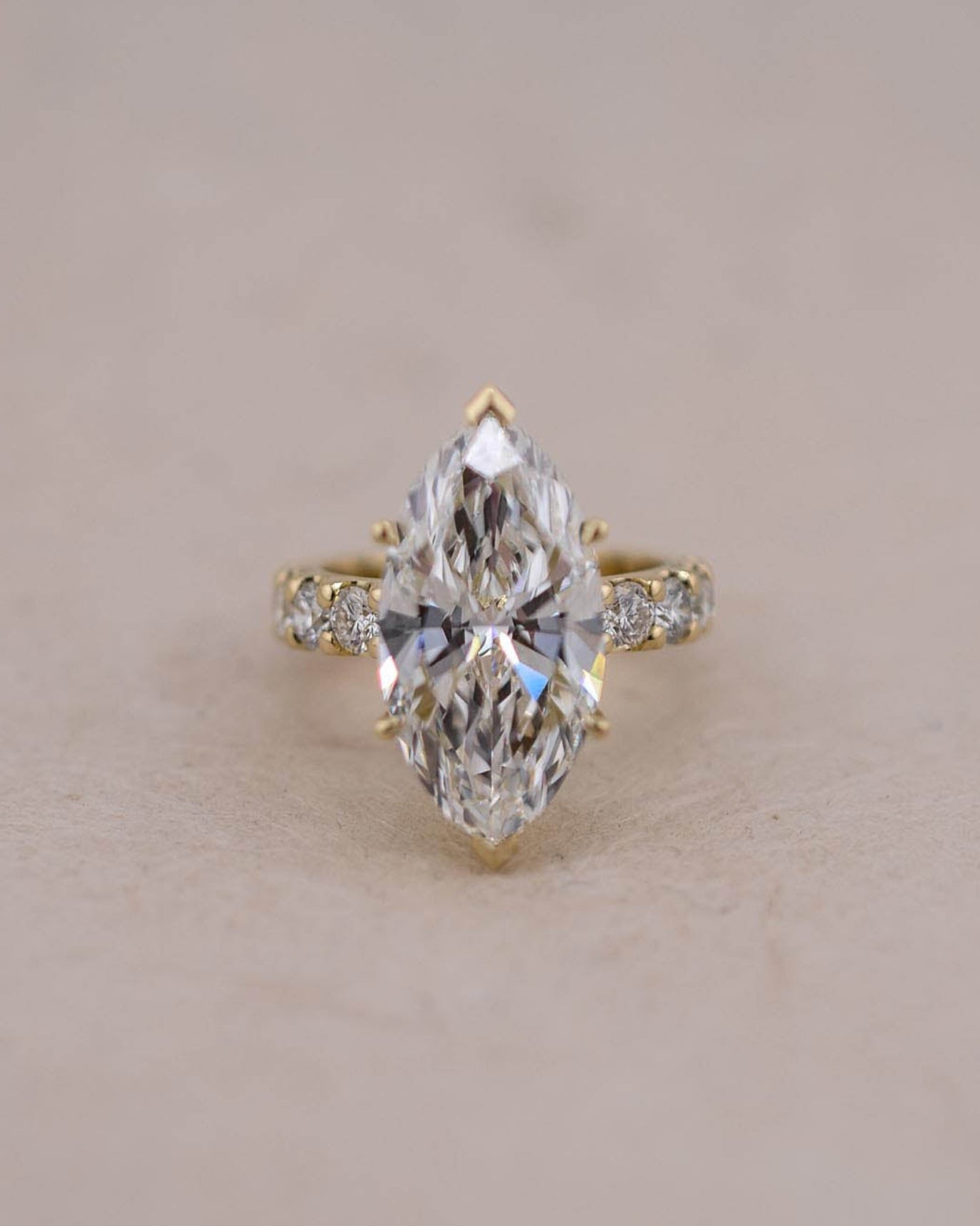A yellow gold ring featuring a marquise-cut white diamond in a prong setting, accented with small round diamonds on the band, set against a plain beige background.