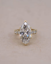 A yellow gold ring featuring a marquise-cut white diamond in a prong setting, accented with small round diamonds on the band, set against a plain beige background.