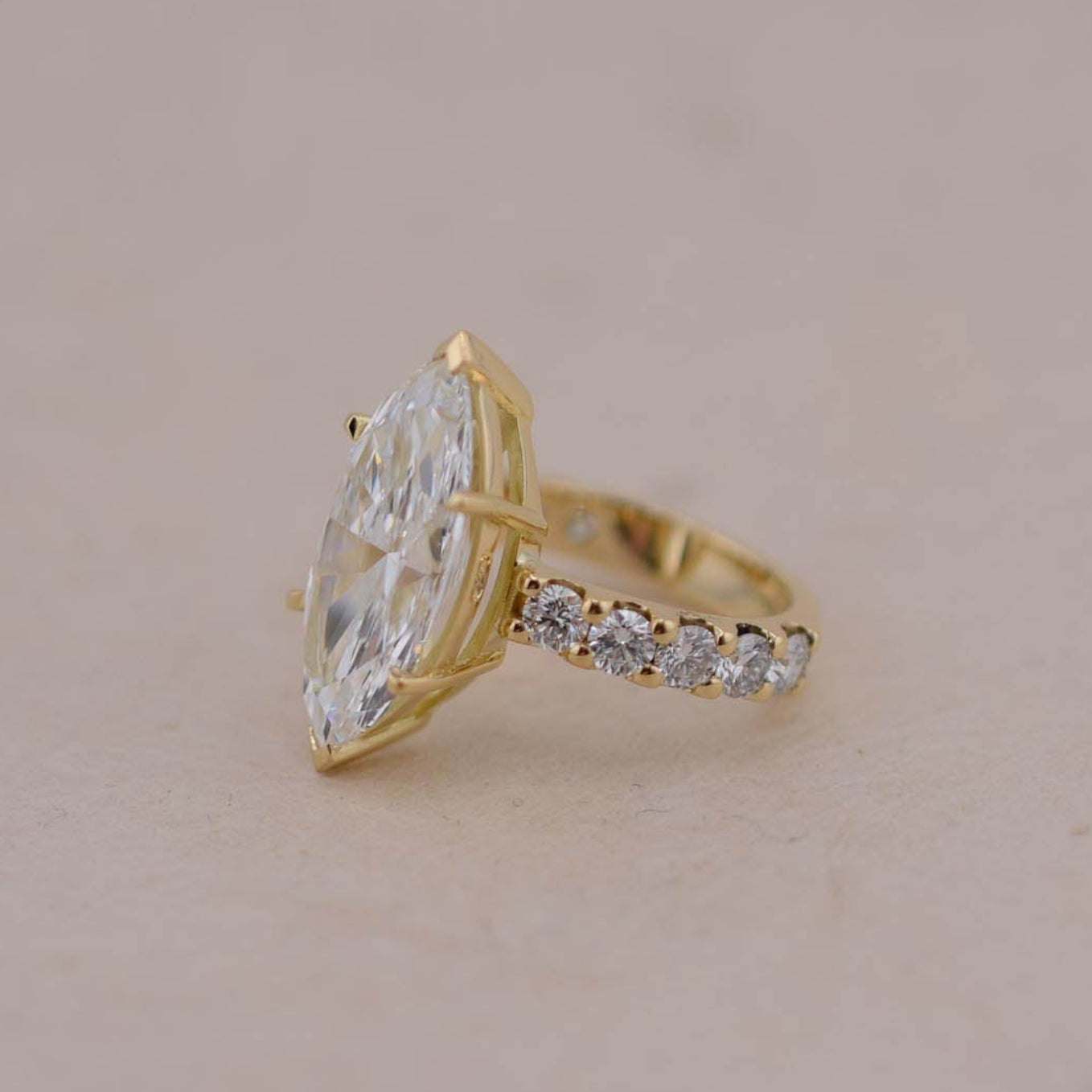 A gold ring featuring a large marquise-cut white diamond and a pavé band with small clear stones, set against a plain beige background.