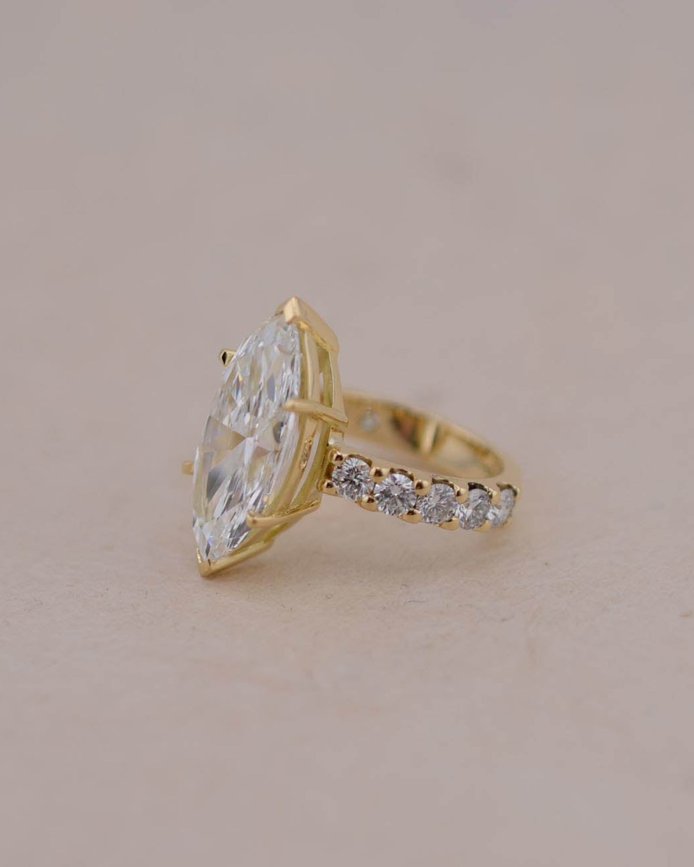 A gold ring featuring a large marquise-cut white diamond and a pavé band with small clear stones, set against a plain beige background.