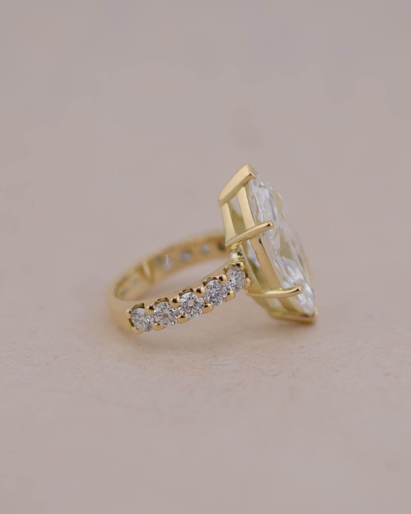 A yellow gold ring featuring a large marquise-cut white diamond and a pavé band with small clear stones, set against a solid light beige background.