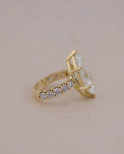 A yellow gold ring featuring a large marquise-cut white diamond and a pavé band with small clear stones, set against a solid light beige background.